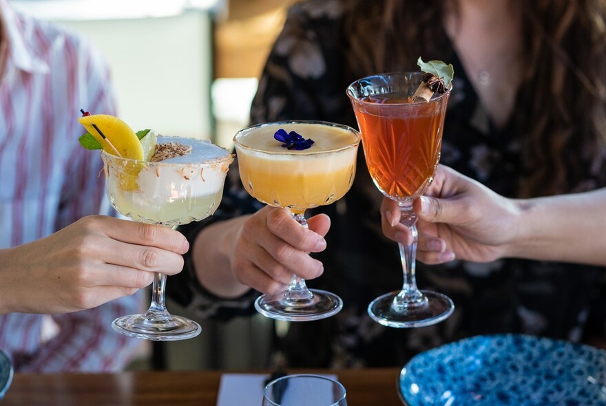 Three people clinking glasses filled with colourful cocktails. 