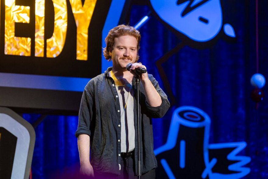 A smiling young man standing on a stage, talking into a microphone on a stand.