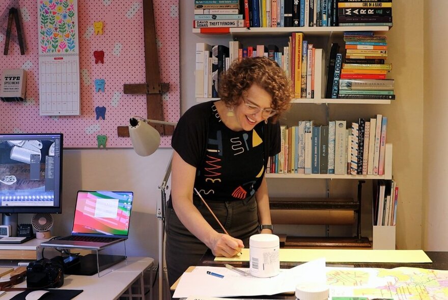 An artist standing, working at a desk with bookshelves and computers in the background. 