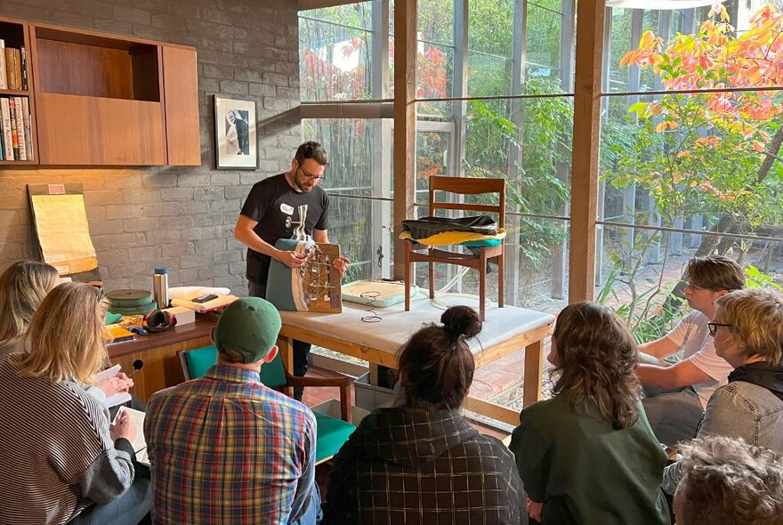 Edwin Fox demonstrating a furniture restoration technique in front of a seated audience in a mid-century room with exposed brick and windows.