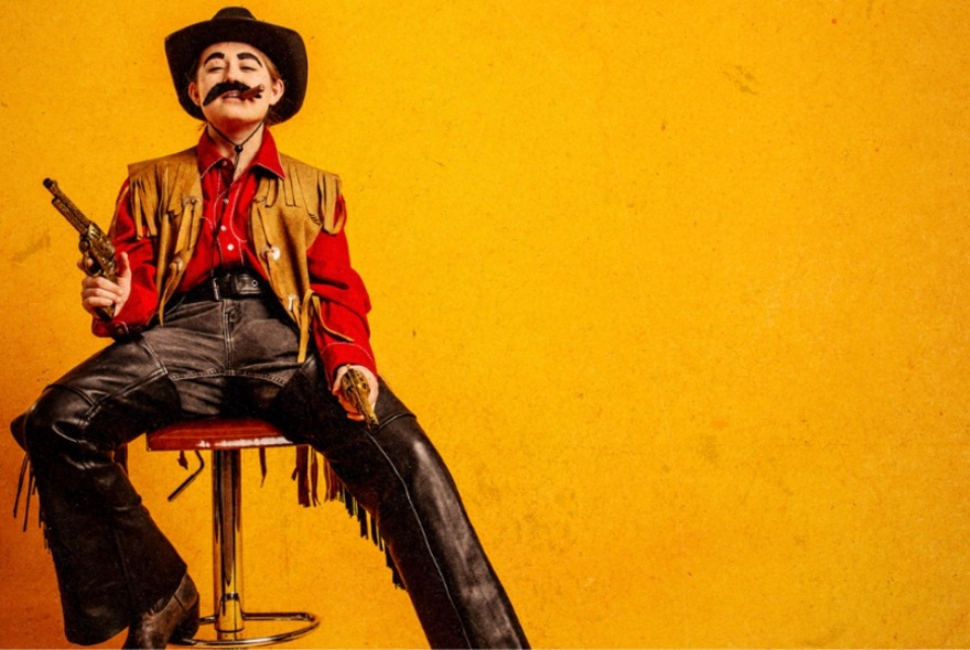 Comedian, Lil Wenker, dressed as a Wild West cowboy, posing on a stool with one pistol raised.