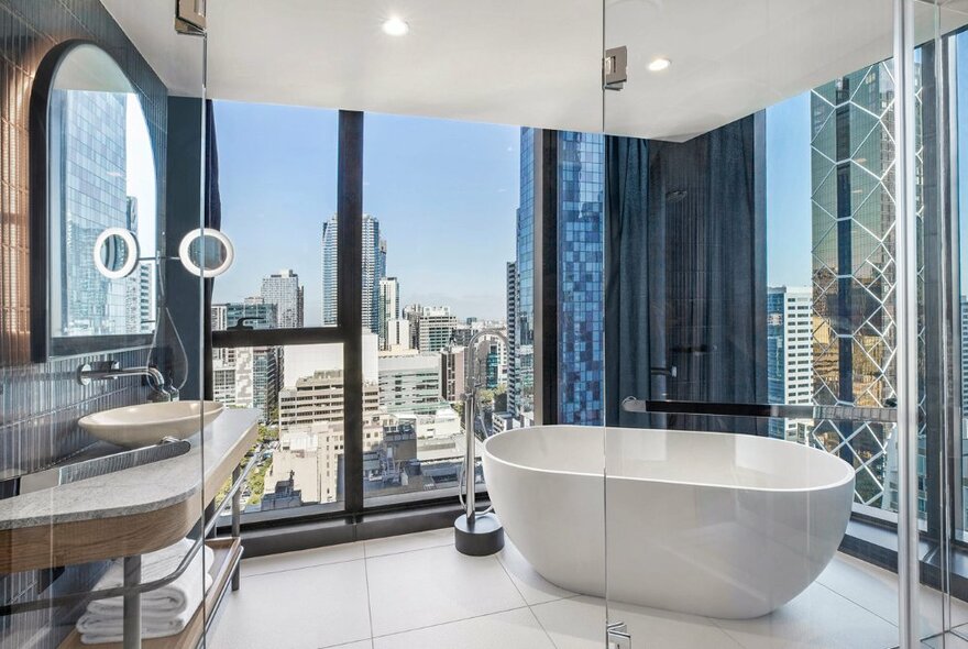 Bathroom at a hotel room in Voco Melbourne Central, with a white bathtub positioned in front of large floor to ceiling windows with panoramic views of the city.