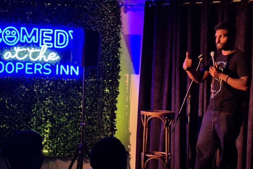 Male stand-up comedian talking into a microphone and standing on a stage, neon signage of club's name visible on the left of the image.