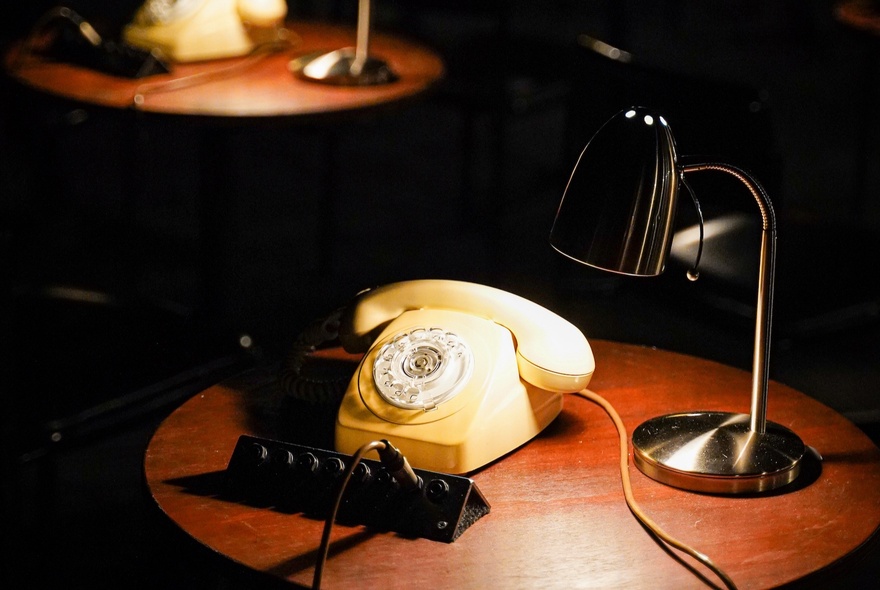 A yellow vintage phone on a wooden table illuminated by a desk lamp, part of an interactive theatre installation at the Malthouse Theatre.