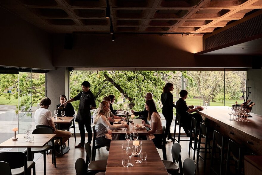 An elegant and moody wine bar with patrons seated at wooden tables, waiters pouring wine and two people at a high bench table. Leafy trees seen through the window.