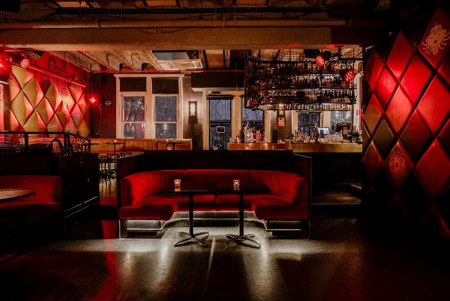 Tables and chairs and booth seating at the bar The Toff in Town, with large windows behind the bar, a reddish glow to the space, and large windows behind the bar area at the rear.