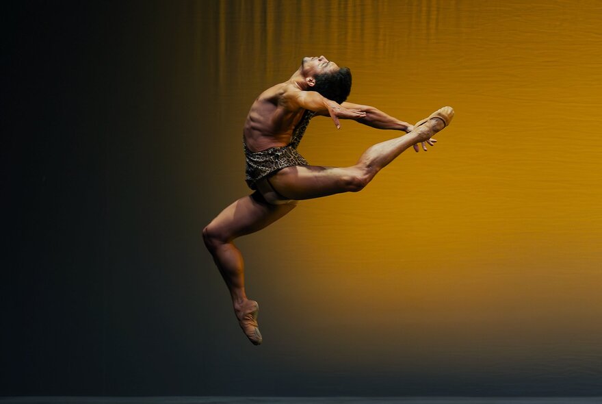 Male dancer pivoting on one toe with arms and leg stretched backwards in an amber light.