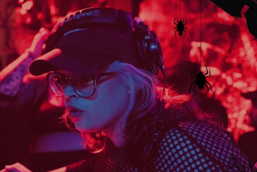 A person with blonde hair, wearing a black cap, glasses, and headphones, DJing under red stage lighting with decorative plastic spiders hanging around them.