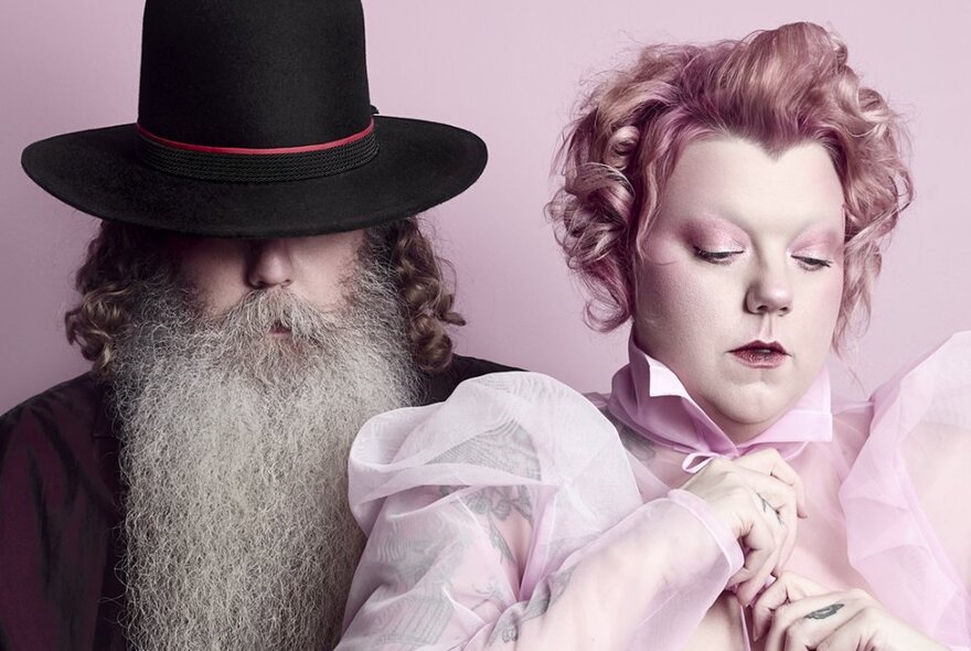 Man with a woolly white beard wearing a dark had that covers his eyes, standing next to a woman with a pale face, stylised pink toned hair wearing a puffed-sleeve pale pink chiffon blouse, her eyes looking downwards.