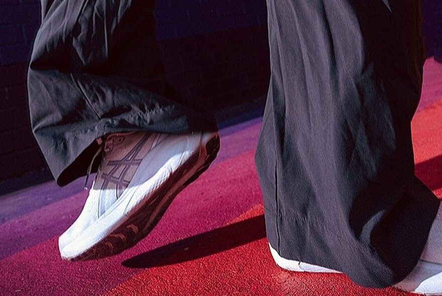 The lower part of a dancer's legs, wearing baggy black pants and Asics runners, with feet facing in opposite directions.