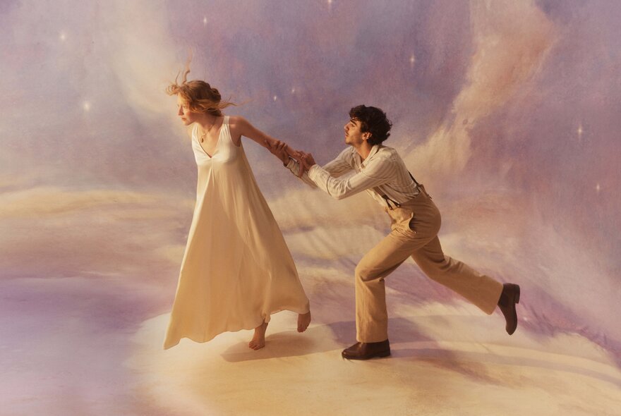 Two performers wearing pale clothing, the man behind a women, whose arm he is hanging onto, against a cloud like backdrop.