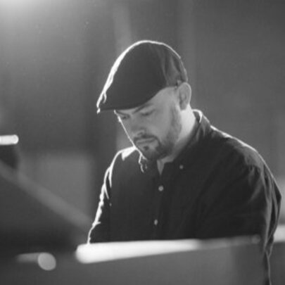 Jazz musician Jack Earle seated at a piano looking downwards wearing a cloth cap. 