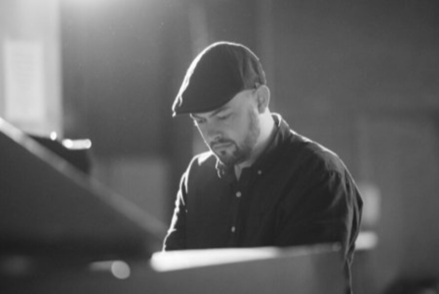 Jazz musician Jack Earle seated at a piano looking downwards wearing a cloth cap. 