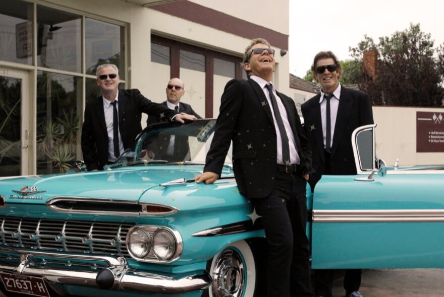 Four men wearing dark suits, ties and sunglasses, posing with turquoise vintage 1950s car.