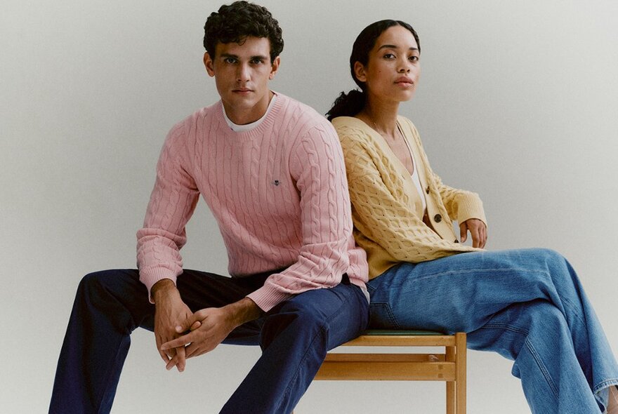 Two models in pastel cable knits and jeans seated casually on one chair. 