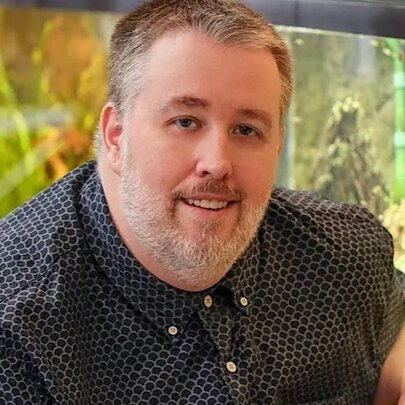 Large man with grey beard, seated in front of an illuminated fish tank.