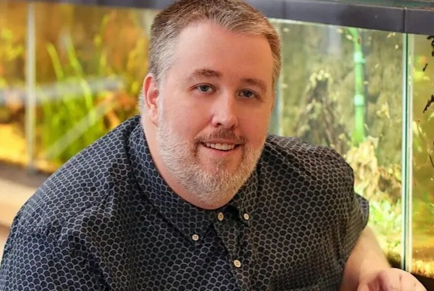 Large man with grey beard, seated in front of an illuminated fish tank.
