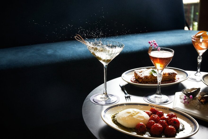 Plates of food and cocktails on a small table, a velvet couch in the background, inside a nightclub.