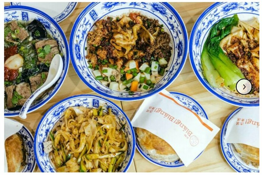 Looking down on a table with bowls of assorted noodles and pastries in paper bags.