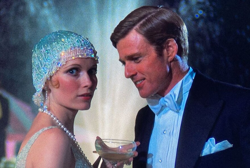 A still from a movie with a couple in 1920s formal wear, the woman wearing a jewelled head covering and long pearls.