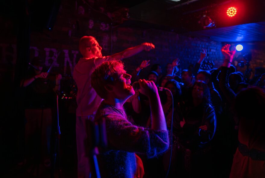 A dark performance space lit with red and blue lights, two people performing. 