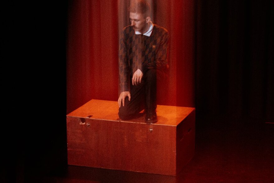 A dark and blurry image of a man crouching on a wooden box.