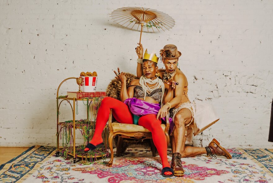A person wearing a gold crown and bright red tights sitting on a vintage armchair, giving a peace sign. Another person in a top hat and a light-coloured outfit is sitting next to them, holding a white parasol over both of them. They are in a room with a white textured wall and a patterned rug.