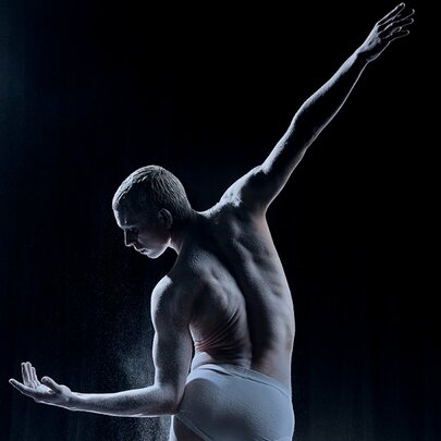 View of a dancer from behind with a bare torso, leaning over sideways to look at his hand on a dark stage with a single spotlight. 