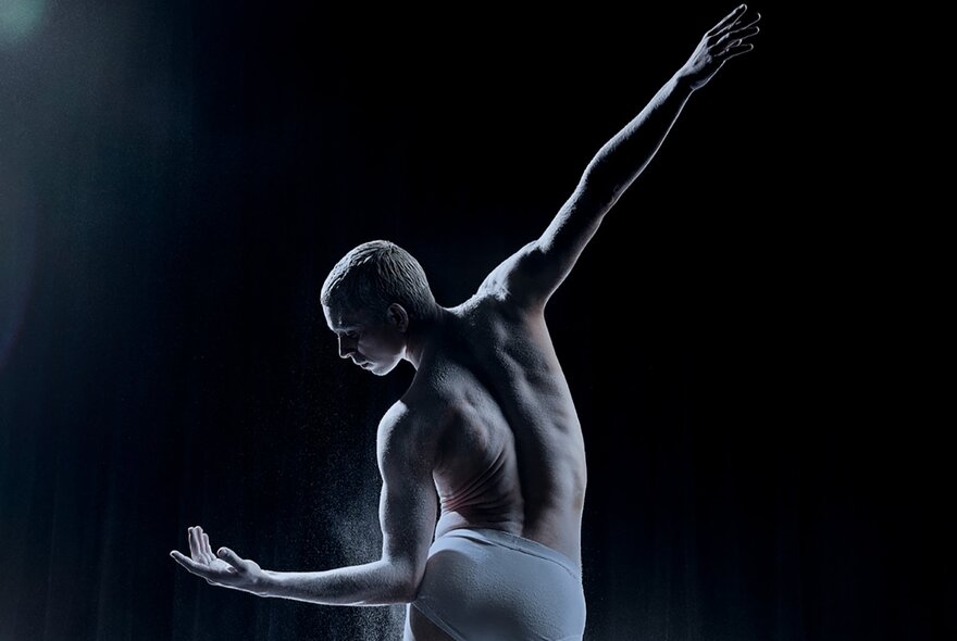 View of a dancer from behind with a bare torso, leaning over sideways to look at his hand on a dark stage with a single spotlight. 