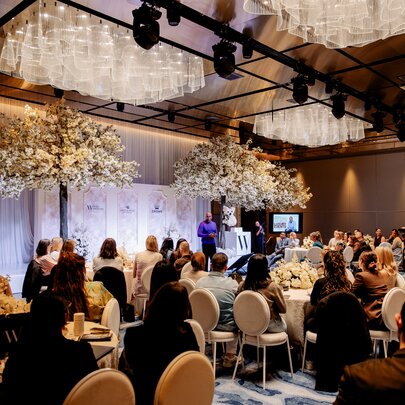 A crowded luxury ballroom event with white floral decor trees and round table seating.