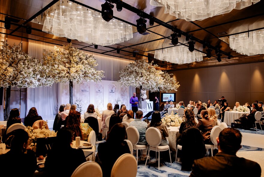 A crowded luxury ballroom event with white floral decor trees and round table seating.