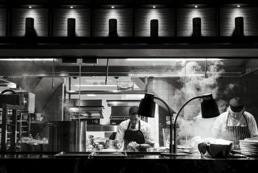Chefs working in a busy commercial restaurant kitchen with steam and counter lighting.