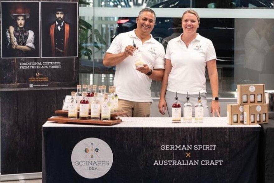 Two people standing behind a stall that is promoting and displaying bottles of flavoured Schnapps.