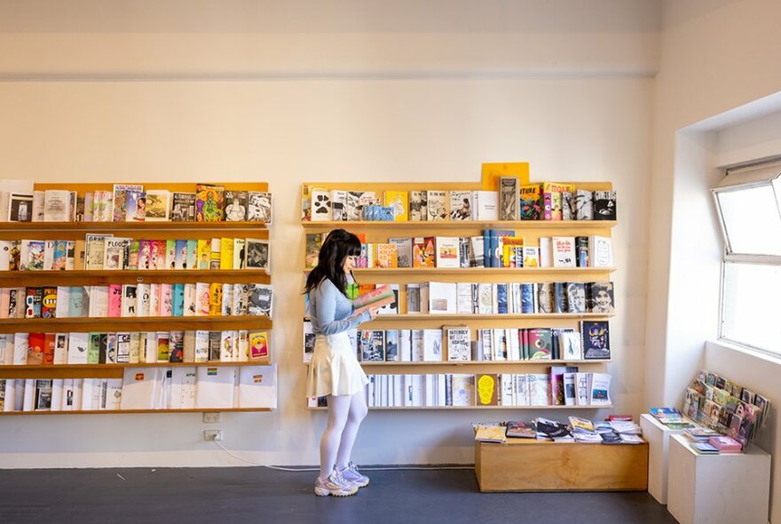 A woman reading a zine in a store with yellow shelves filled with zines.