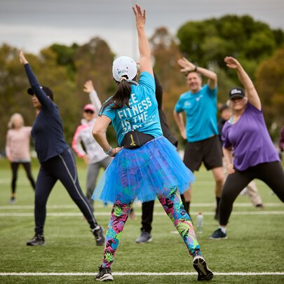 A woman in a blue tutu and bright leggings leads an outdoor group fitness class on a grassy field.
