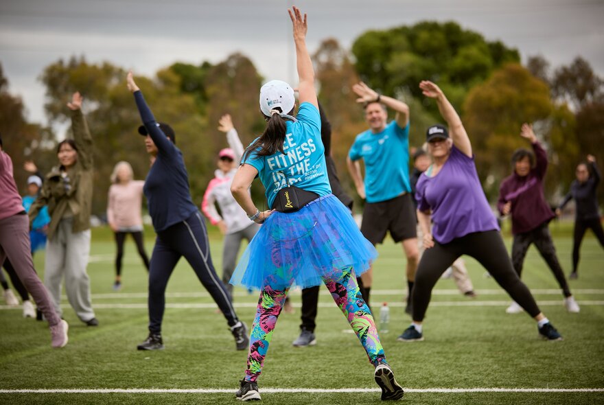 A woman in a blue tutu and bright leggings leads an outdoor group fitness class on a grassy field.
