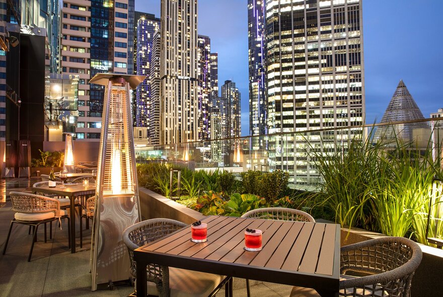 Small tables and chairs on an outdoor terrace at a restaurant with views of Melbourne city buildings and skyline at night.