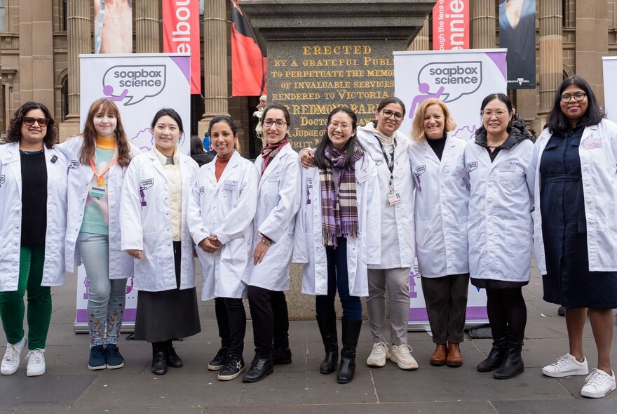 A row of women wearing white coats, standing in front of banners.