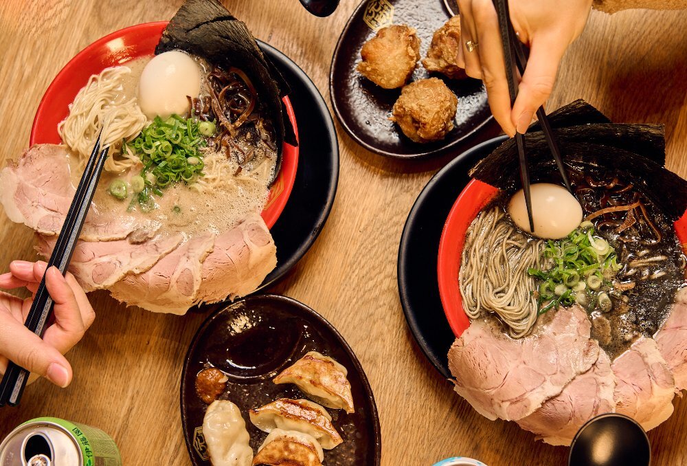 Aerial view of two large bowls of ramen, a side dish of karaage and gyozas and a canned drink.