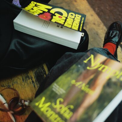 An angled shot shows two books resting on people's laps as they sit side-by-side on a wooden seat.