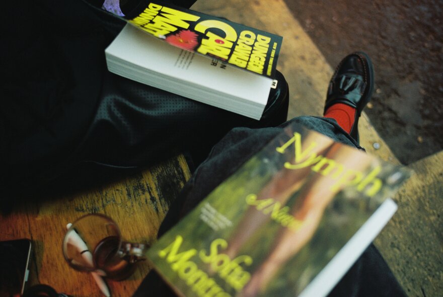 An angled shot shows two books resting on people's laps as they sit side-by-side on a wooden seat.