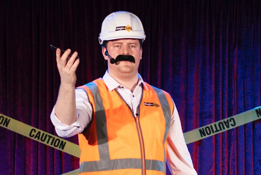 A person wearing a white hard hat, an orange reflective vest, and a fake mustache, performing on a stage.