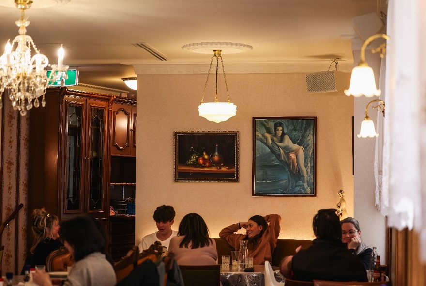 The interior of a restaurant that looks like a lounge room with kitsch artworks on the wall, bookcases, chandeliers, and lace curtains.