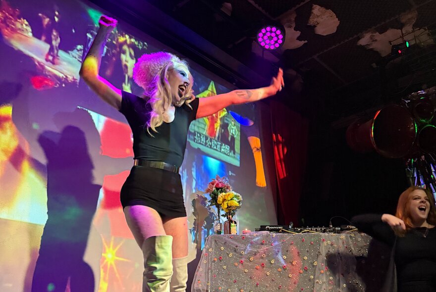 A drag performer on a small stage wearing a black t-shirt, shorts and knee high boots, illuminated with coloured spotlights.