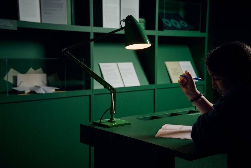 A green room, with a person seated at a green desk writing with a pen on paper, under a green lamp.