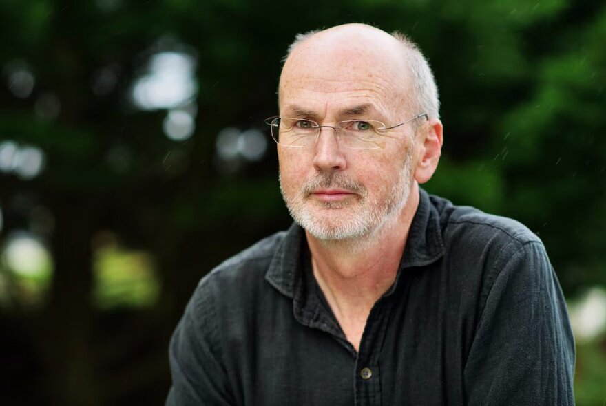 Author Niall Williams, a middle aged balding man with a closely cropped greying beard, wearing glasses and with a neutral expression on his face.