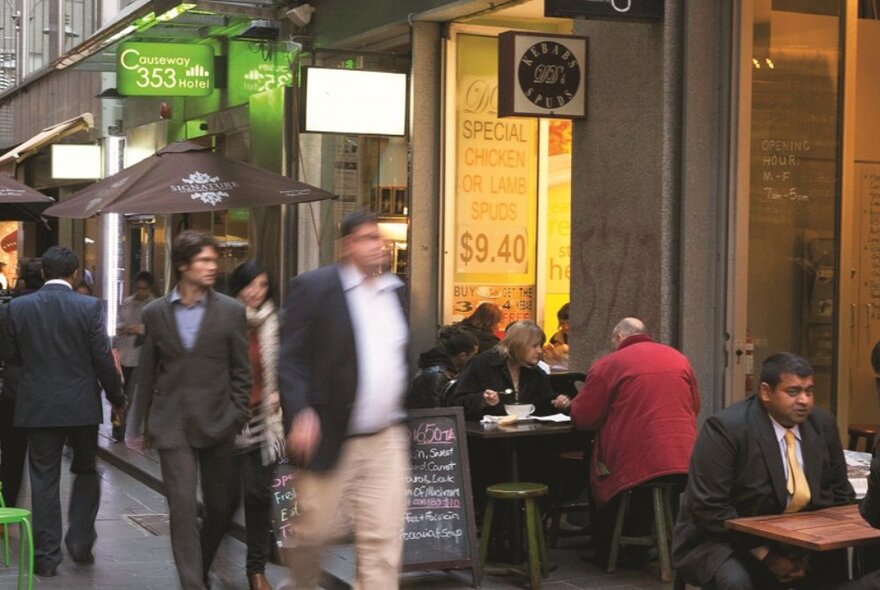 People walking down a busy city laneway with restaurant and hotel signage and cafe tables.