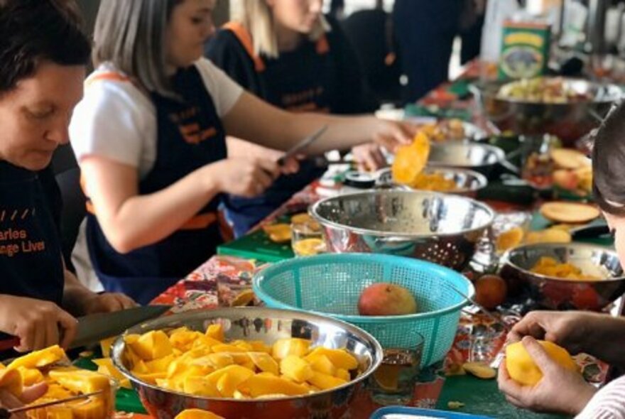 Cooks preparing food seated at a long table, with multiple silver and blue bowls holding mango and other fruits.