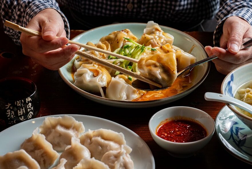 Hands selecting a dumpling with chopsticks and a spoon from a bowl of dumplings in broth.