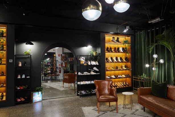 A dimly lit shoe store with black walls, illuminated shelves and tan leather furniture.